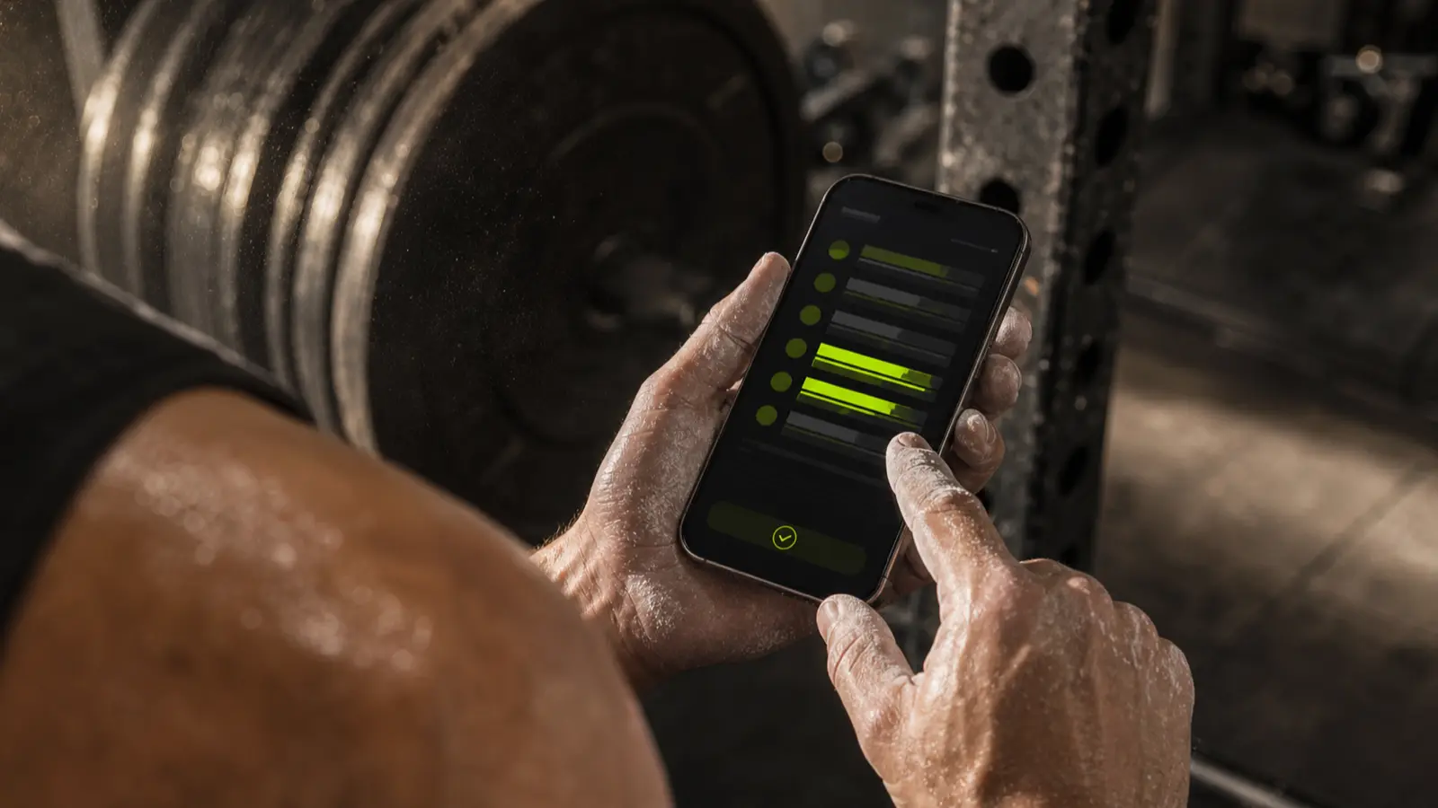 A lifter checking a set on their phone between reps in a dark gym, chalked hands, the workout log glowing volt green